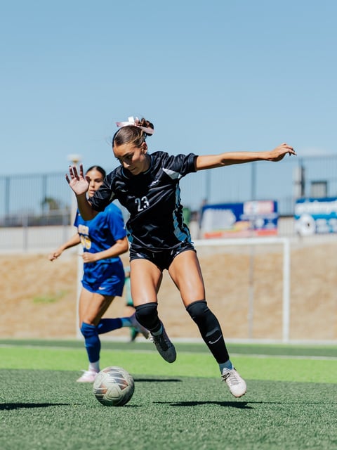 Female football player in black jersey dribbling ball during match with opponent in blue jersey pursuing