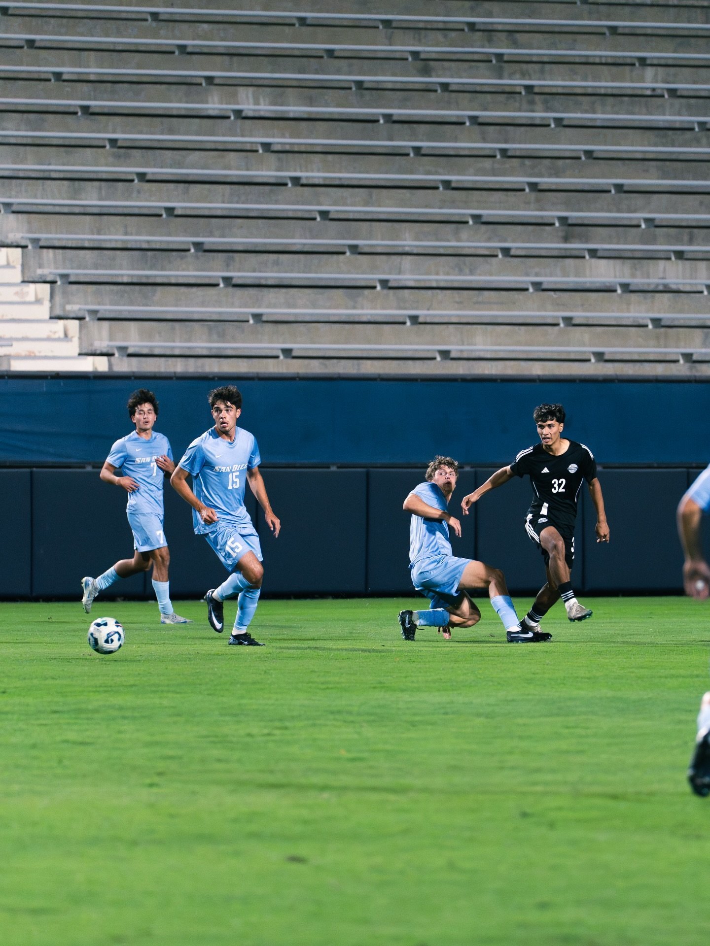 Soccer match with players in light blue and black jerseys competing on a field in front of stadium bleachers
