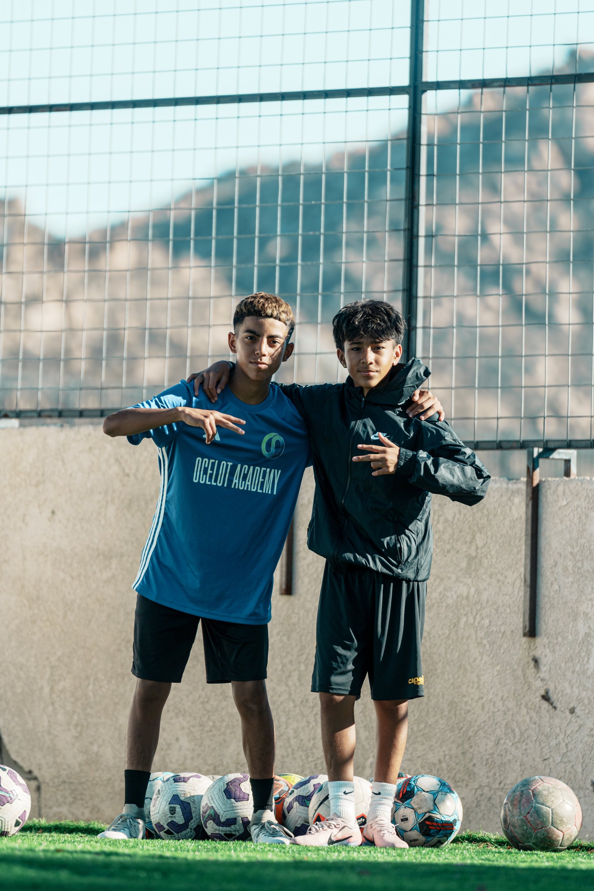 Two young soccer players posing together in front of a goal net with soccer balls lined up below them