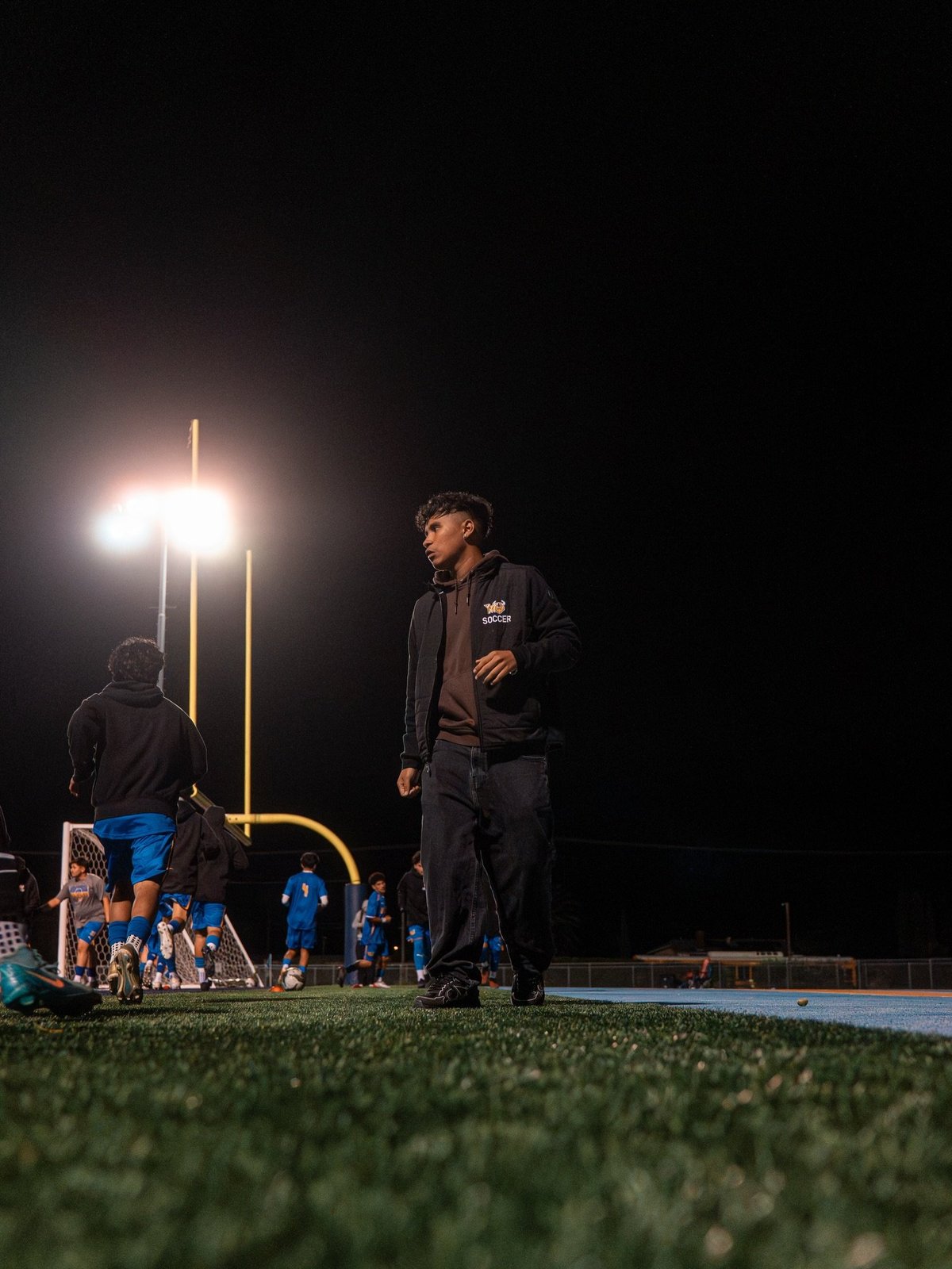 Coach standing on a nighttime football field under bright stadium lights with players in the background