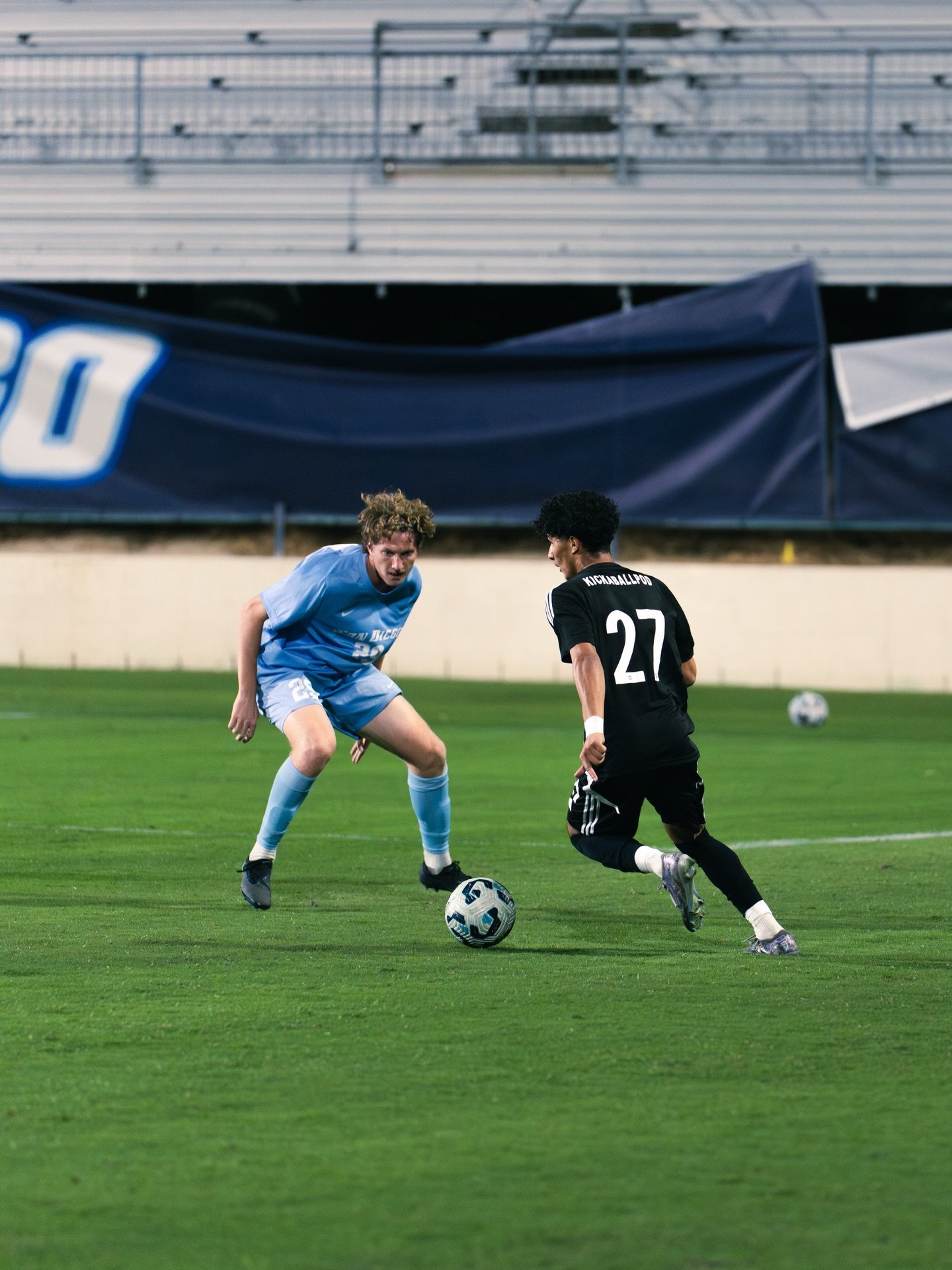 Soccer player in blue jersey dribbles ball while being defended by player in black jersey number 27 on stadium field