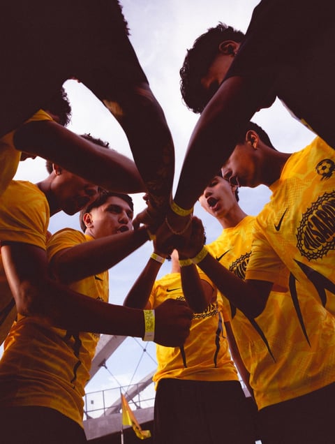 Team members in yellow shirts huddle together with arms joined in a circle, viewed from below against the sky
