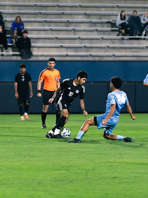 Soccer player in black jersey dribbling ball while opponent in blue jersey attempts to defend during match in stadium