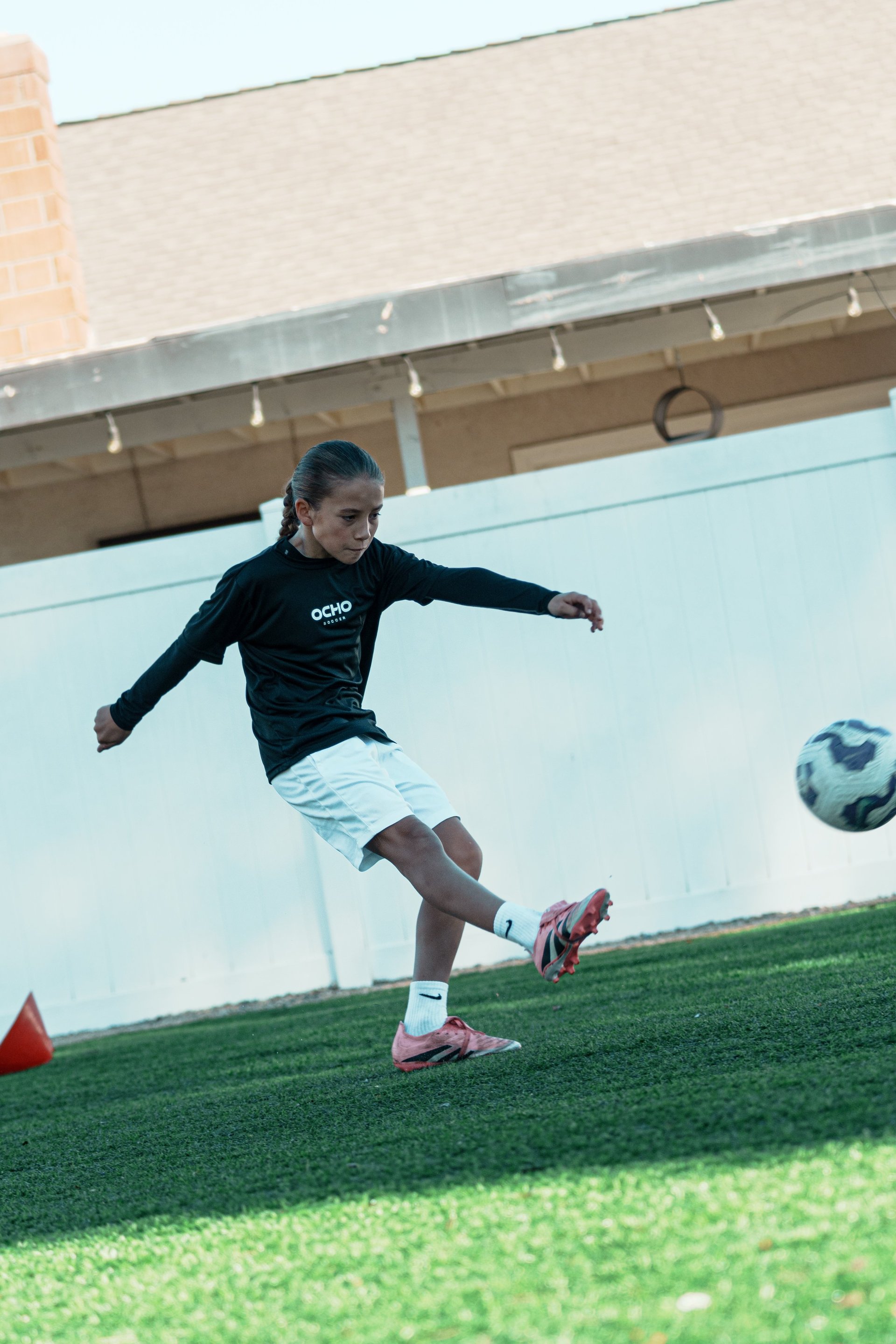 Young female soccer player kicking a ball on artificial turf field with covered structure in background