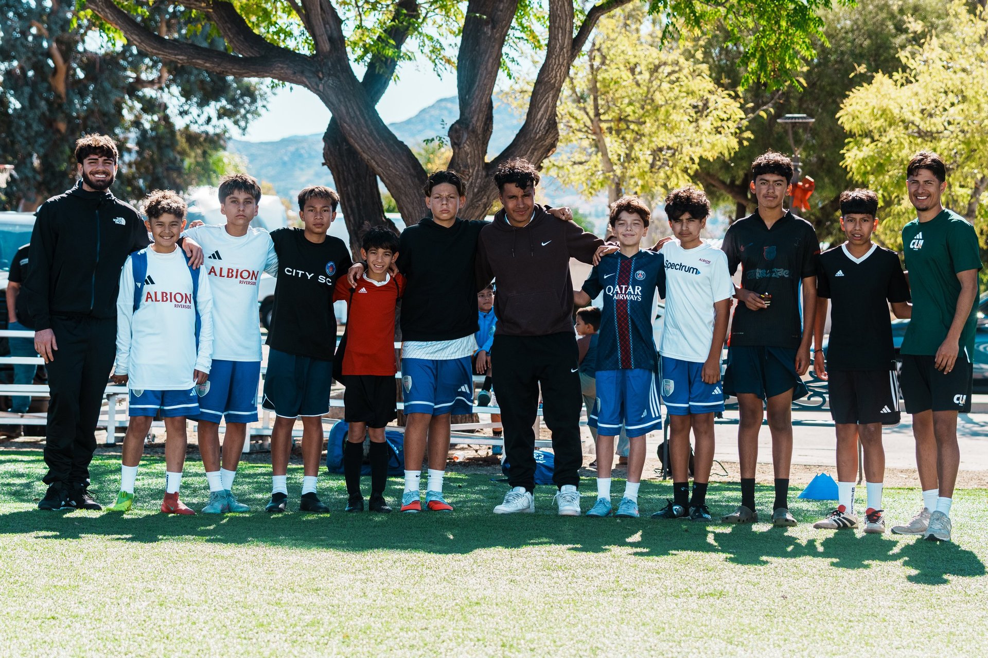 Group of young soccer players and coaches standing together under a large tree on a grass field at a park